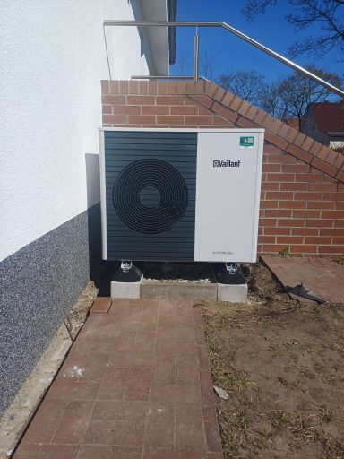 Wärmepumpe an der Wand neben einer Treppe, mit blauem Himmel im Hintergrund.