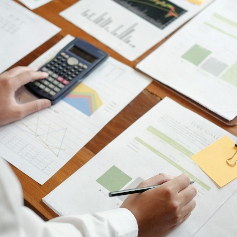 Hände mit Taschenrechner und Stift auf Diagramme und Statistiken auf einem Tisch.